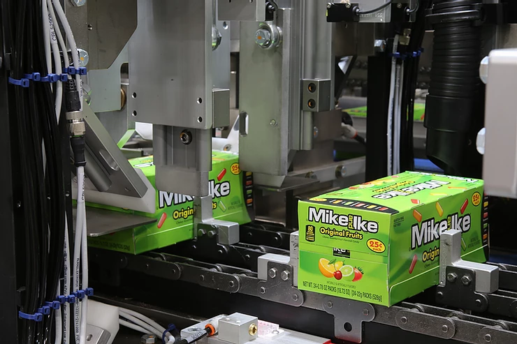 Green Mike and Ike candy boxes on an assembly line in a factory. Boxes have fruit images and are being processed by machinery.