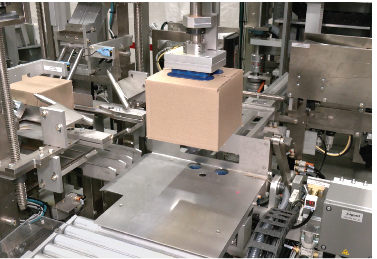 A robotic arm lifts a cardboard box in an industrial machine setting, surrounded by metal components and conveyor belts.