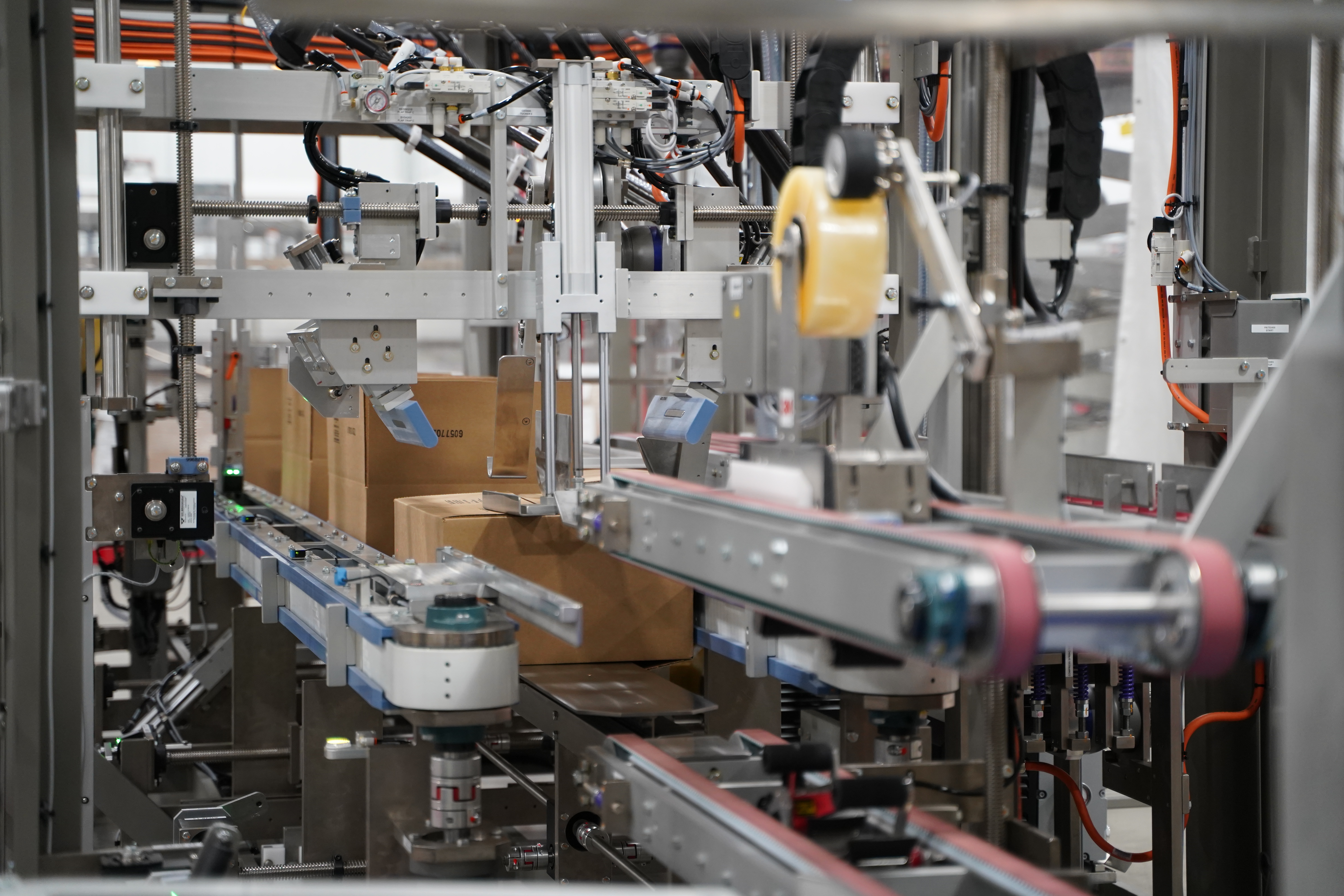Automated packaging machine with conveyor belts moving cardboard boxes in a factory setting, featuring metallic parts and orange cabling.